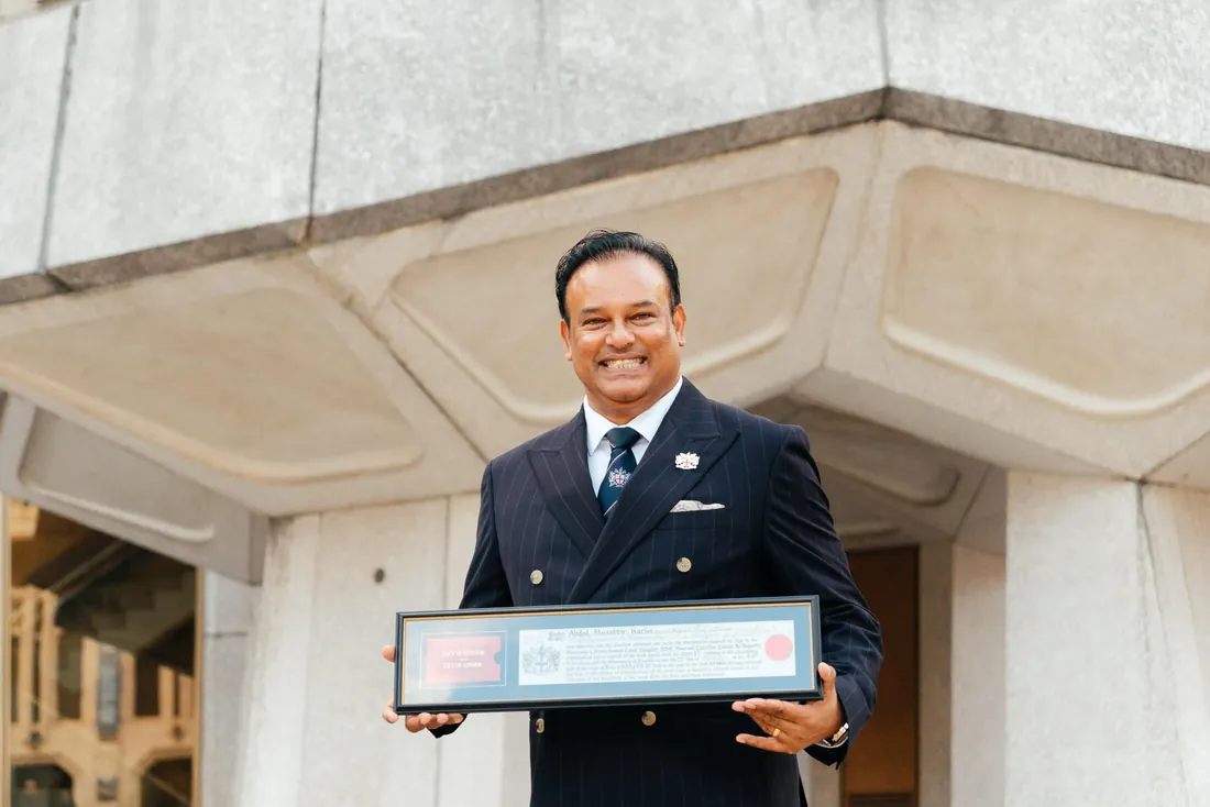 Sabbir Karim smiling outside a stone building, holding his framed copy of the Freedom of the City of London