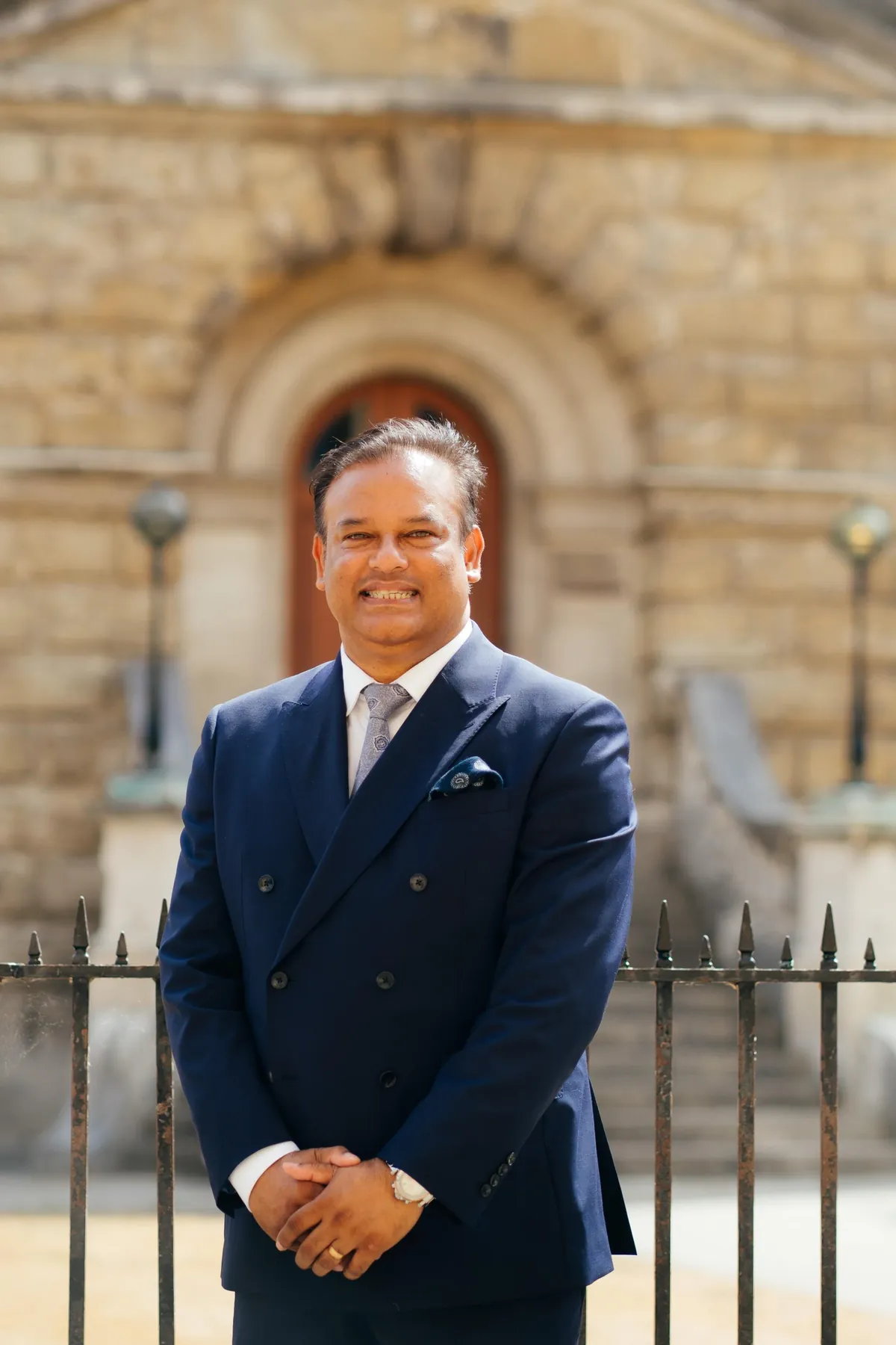 Sabbir Karim in a navy double-breasted suit, standing by iron railings in front of a stone building